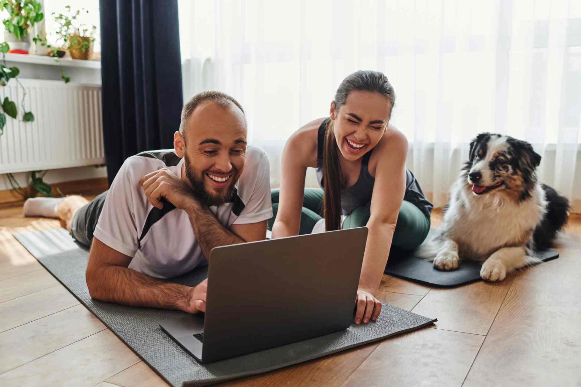 Joyful couple in sportswear using laptop together on fitness mats near border collie at home